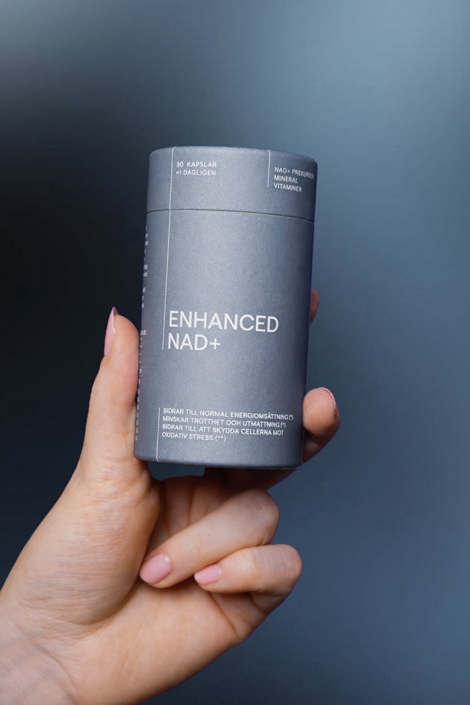 En hand med ljusrosa nagellack håller i en mörkgrå KÄLLA ENHANCED NAD+ behållare mot en blågrå bakgrund. Etiketten lyfter fram vitaminer, cellulär energi och fördelar som stödjer ett hälsosamt åldrande.